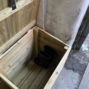 May include: A wooden storage box with a hinged lid, open to reveal a pair of black rubber boots inside. The box is made of unfinished wood and has a simple design.