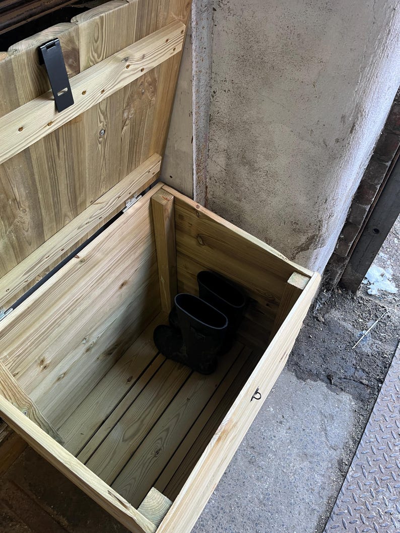 May include: A wooden storage box with a hinged lid, open to reveal a pair of black rubber boots inside. The box is made of unfinished wood and has a simple design.