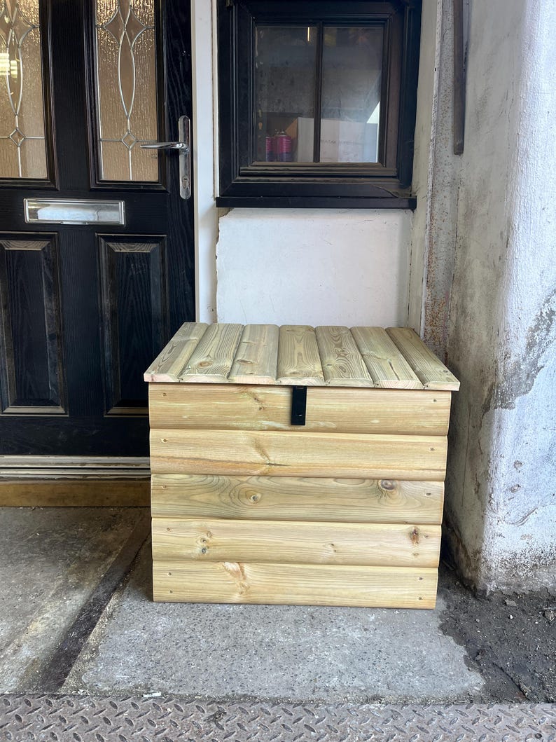 May include: A wooden storage box with a hinged lid and a black metal latch. The box is made of natural wood and has a light brown color.