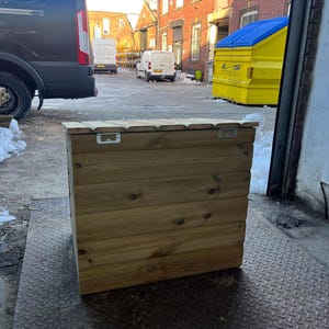 May include: A wooden storage box with a hinged lid. The box is made of natural wood and has a rustic look. The box is sitting on a metal grate.