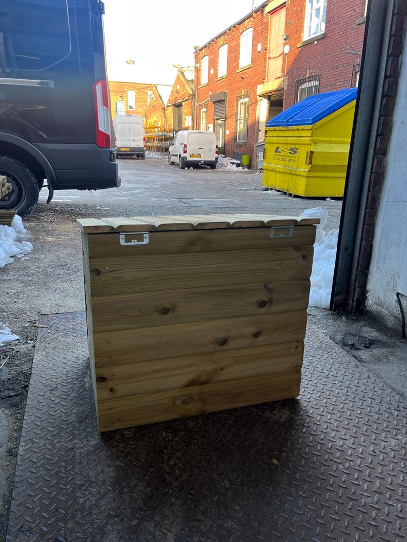 May include: A wooden storage box with a hinged lid. The box is made of natural wood and has a rustic look. The box is sitting on a metal grate.