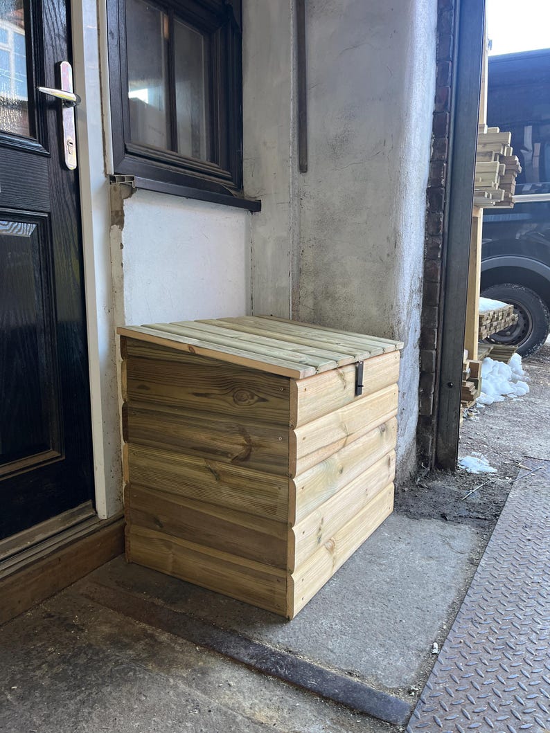 May include: A wooden storage box with a hinged lid, made of natural wood with a light brown finish. The box is sitting on a concrete surface next to a door.