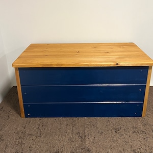 May include: A wooden storage chest with a natural wood top and a dark blue painted body. The chest has three horizontal panels on the front.