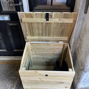 May include: A wooden storage box with a hinged lid, made of light brown wood with a natural finish. The box is open and shows the interior, which is empty except for two black rubber boots.