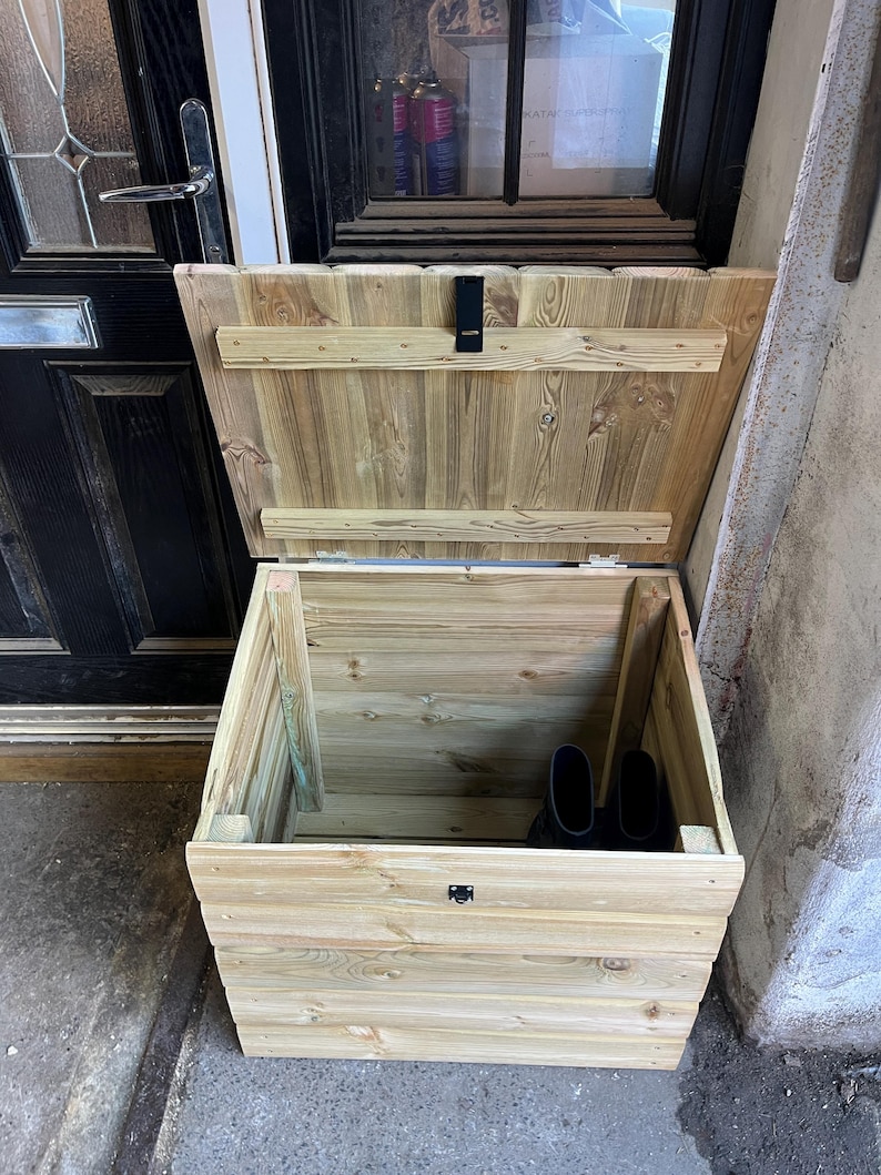 May include: A wooden storage box with a hinged lid, made of light brown wood with a natural finish. The box is open and shows the interior, which is empty except for two black rubber boots.