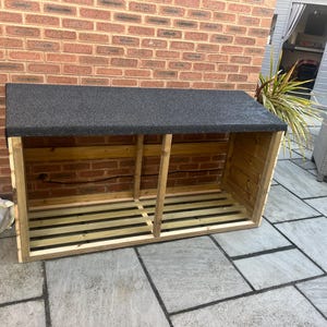 May include: A wooden log store with a dark grey, textured roof. The unit has two sections and is made from natural wood. The structure is outdoors, positioned against a brick wall and a paved patio.