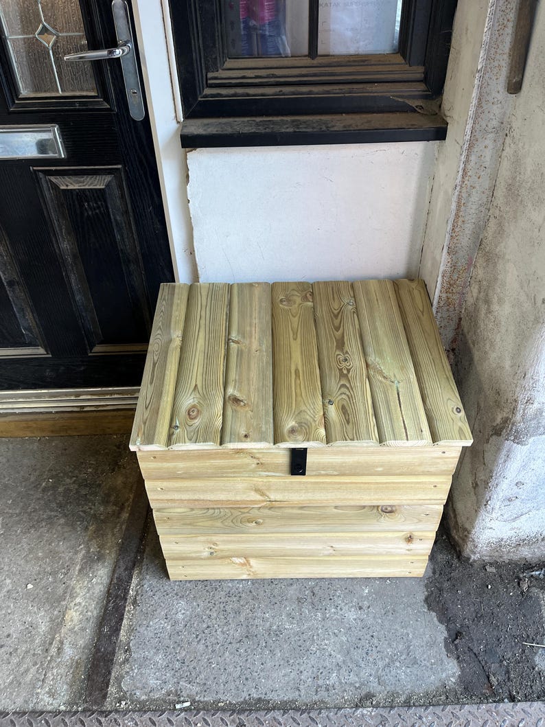 May include: A wooden storage box with a hinged lid and a black metal latch. The box is made of light brown wood and has a natural finish.