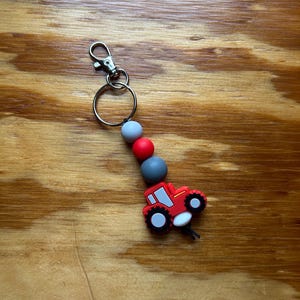 Può includere: Un portachiavi a forma di trattore rosso con moschettone e portachiavi argentati. Il portachiavi presenta tre perline in silicone grigie e rosse. Il trattore ha ruote bianche e nere e un finestrino bianco.