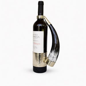 Puede incluir: Una botella de vino con un soporte decorativo en forma de cuerno. La botella es oscura con una etiqueta blanca. El soporte es de metal color oro y un cuerno blanco y negro. El soporte envuelve la botella.