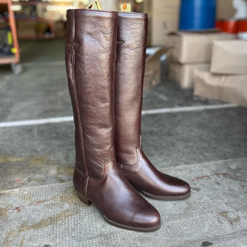 May include: A pair of tall, brown leather riding boots. The boots have a smooth, polished finish and a classic design, with a slight heel. The boots are standing on a concrete surface.