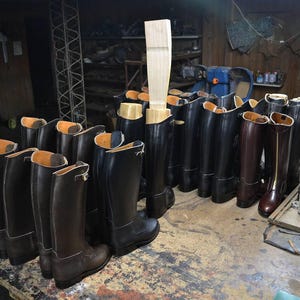 May include: A collection of black leather riding boots with brown leather boots in the front. The boots are lined up in a row on a wooden floor.