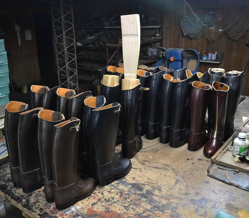 May include: A collection of black leather riding boots with brown leather boots in the front. The boots are lined up in a row on a wooden floor.