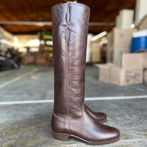 May include: A pair of tall, brown leather riding boots. The boots have a smooth, polished finish and a classic design, with a decorative cross-stitch detail at the top of the shaft. The boots are designed to reach the knee.