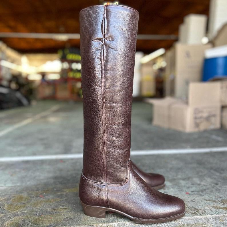 May include: A pair of tall, brown leather riding boots. The boots have a smooth, polished finish and a classic design, with a decorative cross-stitch detail at the top of the shaft. The boots are designed to reach the knee.