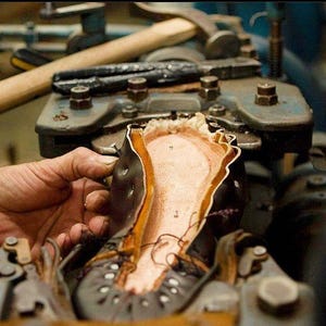 May include: A close-up of a shoe being assembled in a workshop. The shoe is made of leather and has a brown leather sole. The shoe is being held in place by a metal clamp.