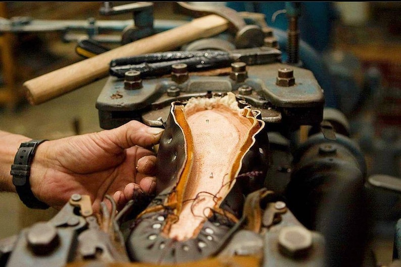 May include: A close-up of a shoe being assembled in a workshop. The shoe is made of leather and has a brown leather sole. The shoe is being held in place by a metal clamp.