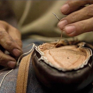 May include: Close-up of a person's hands sewing a brown leather shoe with a needle and thread.