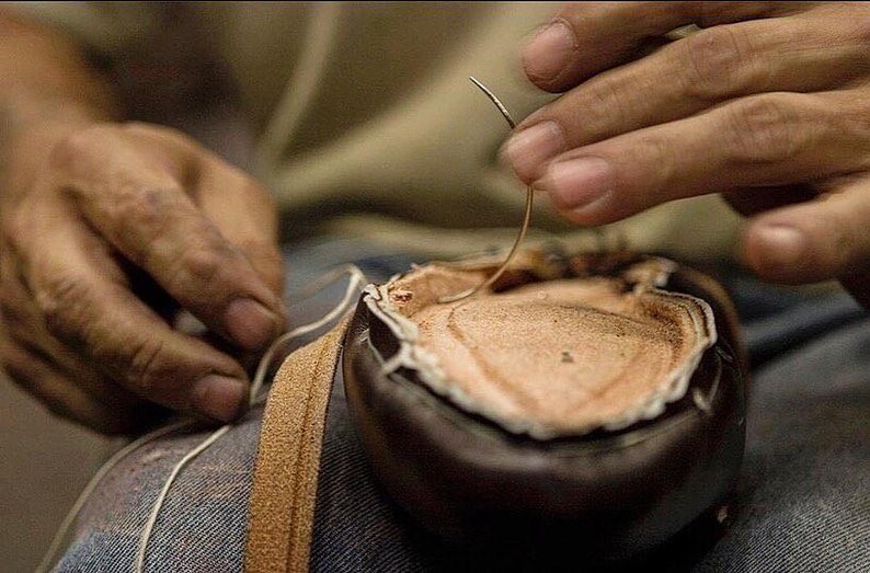 May include: Close-up of a person's hands sewing a brown leather shoe with a needle and thread.