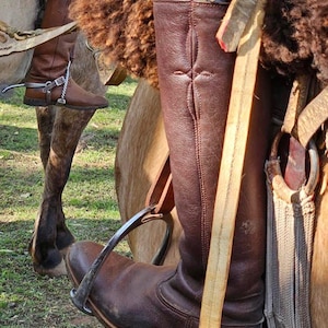 May include: Close-up of brown leather riding boots with spurs and stirrups. The boots are worn by a rider on horseback. The image highlights the equestrian equipment and the texture of the leather.