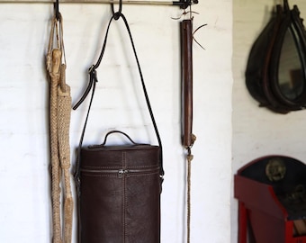 May include: A collection of equestrian equipment hanging on a white brick wall. Items include a brown leather cylindrical bag with a handle, a braided rope, and a wooden riding crop. A wooden rack with metal hooks holds the items.
