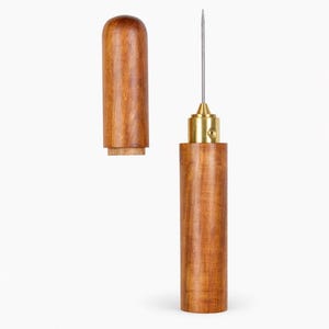 Puede incluir: Una lezna de madera con una férula de latón y una aguja larga y delgada. La lezna está hecha de madera de color marrón claro y tiene forma cilíndrica. La tapa está separada, revelando el interior de corcho. Esta herramienta se utiliza para perforar agujeros en cuero o tela.