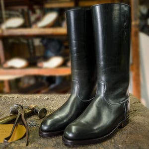 May include: A pair of black leather riding boots with a tall shaft and a pointed toe. The boots are sitting on a wooden surface.