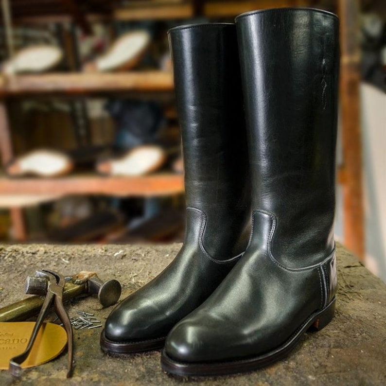 May include: A pair of black leather riding boots with a tall shaft and a pointed toe. The boots are sitting on a wooden surface.