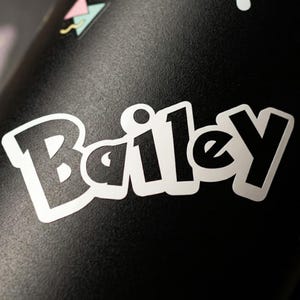 Könnte beinhalten: Ein mattschwarzer Becher mit einem weißen Aufkleber, der den Namen Bailey in einer fetten, abgerundeten Schrift zeigt. Die Oberfläche ist mit kleinen, bunten geometrischen Formen wie Dreiecken und Kreisen in Pastellrosa, Türkis und Gelb verziert.