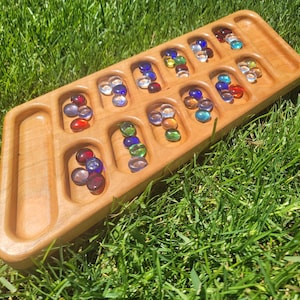 May include: A wooden Mancala board with a light finish, filled with colorful glass marbles. The board has a rectangular shape with rounded pockets and is set on a bed of green grass. The marbles are various colors, including red, blue, green, and yellow.