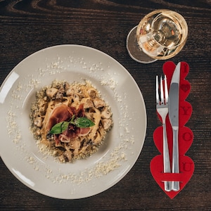 May include: A white plate with a serving of pasta with mushrooms, prosciutto, and a sprig of basil. The pasta is topped with grated cheese. A red heart-shaped place card with the words "I Love You" is on the table next to a fork and knife.