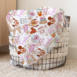 Puede incluir: Una manta de beb&eacute; blanca con un patr&oacute;n colorido de personajes de Disney, incluyendo Mickey Mouse, Minnie Mouse, Donald Duck y Goofy. La manta presenta un dise&ntilde;o rosa y dorado con el nombre "Sophia Ann" repetido en todo.