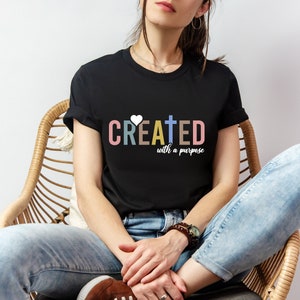 Puede incluir: Camiseta negra de cuello redondo con el texto "CREATED with a purpose" en letras coloridas y un gráfico de corazón y cruz. La persona está sentada en una silla de mimbre, usando jeans azules y zapatos marrones.