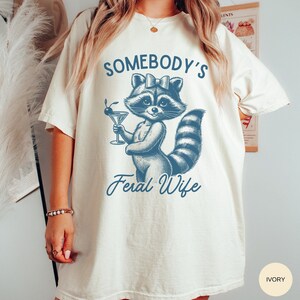 May include: A white t-shirt with a blue illustration of a raccoon holding a martini glass. The text "Somebody's Feral Wife" is printed below the raccoon. The shirt is available in ivory.