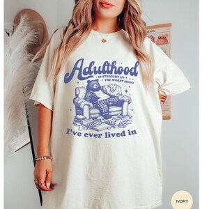 Puede incluir: Una camiseta blanca con un gráfico azul de un oso sentado en una silla. El texto "Adulthood is straight up the worst hood" y "I've ever lived in" está impreso en la camiseta.