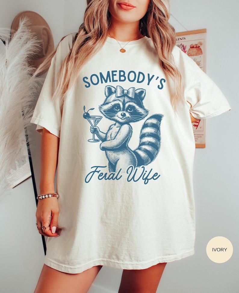 May include: A white t-shirt with a blue illustration of a raccoon holding a martini glass. The text "Somebody's Feral Wife" is printed below the raccoon. The shirt is available in ivory.