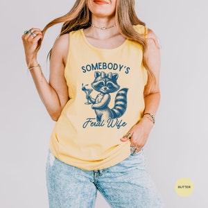 May include: A yellow tank top with a blue graphic of a raccoon wearing a bow and holding a martini glass. The text "Somebody's Feral Wife" is printed below the raccoon.