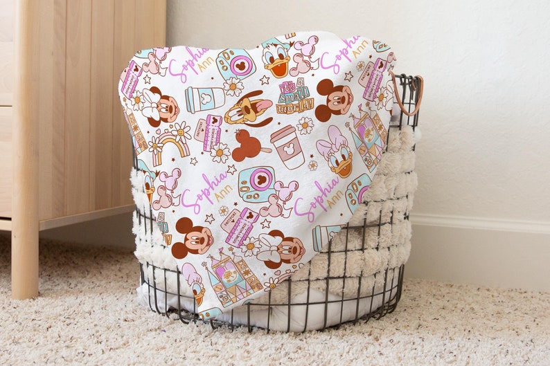 Puede incluir: Una manta de beb&eacute; blanca con un patr&oacute;n colorido de personajes de Disney, incluyendo Mickey Mouse, Minnie Mouse, Donald Duck y Goofy. La manta presenta un dise&ntilde;o rosa y dorado con el nombre "Sophia Ann" repetido en todo.