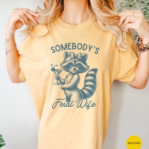 May include: A yellow t-shirt with a blue illustration of a raccoon holding a martini glass. The text "Somebody's Feral Wife" is printed below the raccoon.