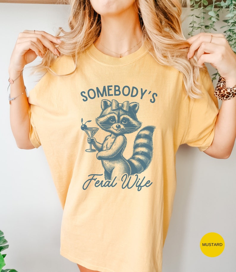 May include: A yellow t-shirt with a blue illustration of a raccoon holding a martini glass. The text "Somebody's Feral Wife" is printed below the raccoon.