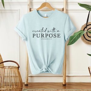 Puede incluir: Camiseta azul claro con el texto "created with a PURPOSE" y "EPHESIANS 2:10" en letras negras.