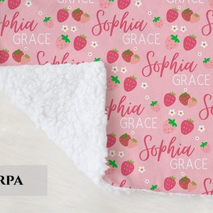 Pode incluir: Uma manta de sherpa rosa e branca com um padr&atilde;o repetido de morangos e o texto "Sophia Grace".