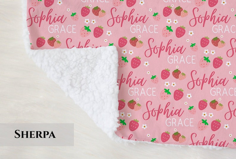 Pode incluir: Uma manta de sherpa rosa e branca com um padr&atilde;o repetido de morangos e o texto "Sophia Grace".