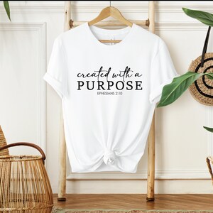 Puede incluir: Camiseta blanca con el texto "created with a PURPOSE" y "EPHESIANS 2:10" impreso en negro.