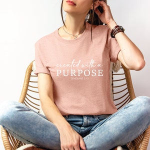 Puede incluir: Una mujer lleva una camiseta rosa con el texto "created with a PURPOSE EPHESIANS 2:10" en letras blancas. Est&aacute; sentada en una silla de mimbre con pantalones vaqueros azules.