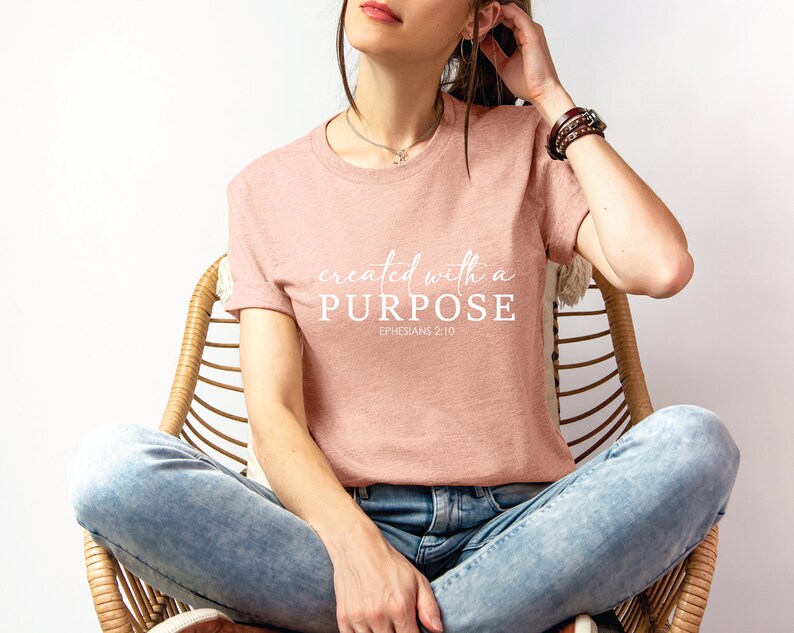 Puede incluir: Una mujer lleva una camiseta rosa con el texto "created with a PURPOSE EPHESIANS 2:10" en letras blancas. Est&aacute; sentada en una silla de mimbre con pantalones vaqueros azules.