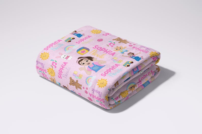 May include: Folded pink blanket with a repeating pattern of cartoon characters, rainbows, and the name "Sophia" in pink and white. The blanket features a variety of illustrations, including a girl, teddy bears, and musical notes.