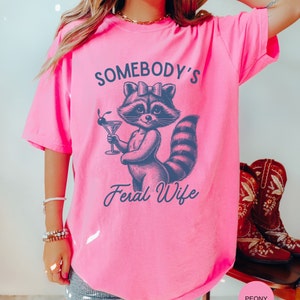 May include: Pink t-shirt with a graphic of a raccoon wearing a bow and holding a martini glass. The text "Somebody's Feral Wife" is printed below the raccoon.