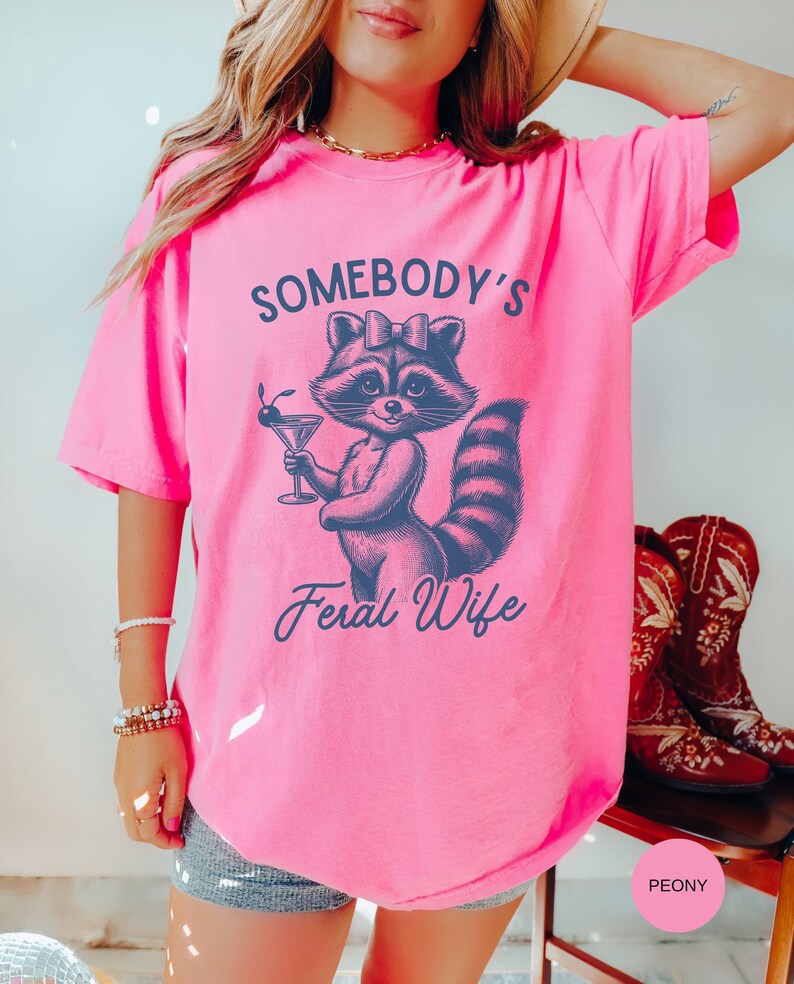 May include: Pink t-shirt with a graphic of a raccoon wearing a bow and holding a martini glass. The text "Somebody's Feral Wife" is printed below the raccoon.