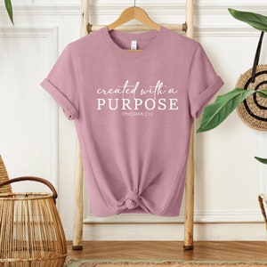 Puede incluir: Una camiseta de color rosa polvoriento con el texto "created with a PURPOSE" y "EPHESIANS 2:10" en blanco.