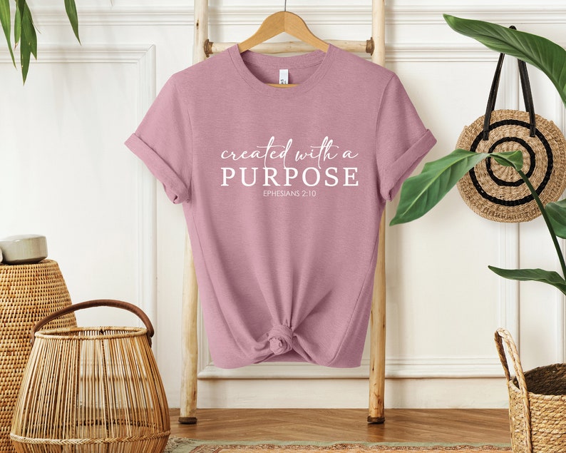 Puede incluir: Una camiseta de color rosa polvoriento con el texto "created with a PURPOSE" y "EPHESIANS 2:10" en blanco.
